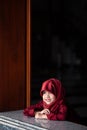 Adorable little muslim girl in traditional clothing, red hijab or niqab and red abaya smiling and watching out the window. Prawet